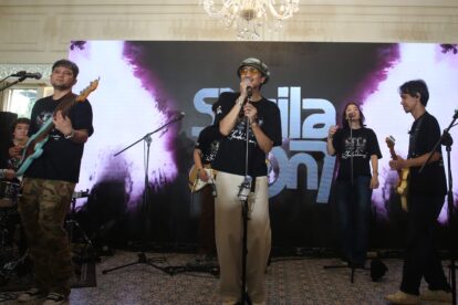 Lead singer wearing a bucket hat and sunglasses sings into a mic on a stage with bandmates playing guitars behind him, chandeliers overhead.