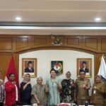 A group of men and women in formal and official attire pose with fists raised in a government meeting room with national symbols on the wall behind them
