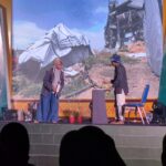 Two people on a stage handling equipment with a mountainous backdrop and stage gear visible in the foreground