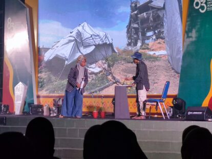 Two people on a stage handling equipment with a mountainous backdrop and stage gear visible in the foreground.