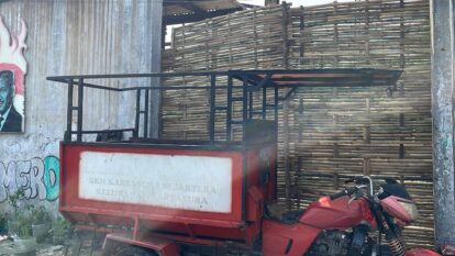 Red trailer with a metal frame and faded white panel parked beside a tall woven bamboo fence and graffiti wall on the left