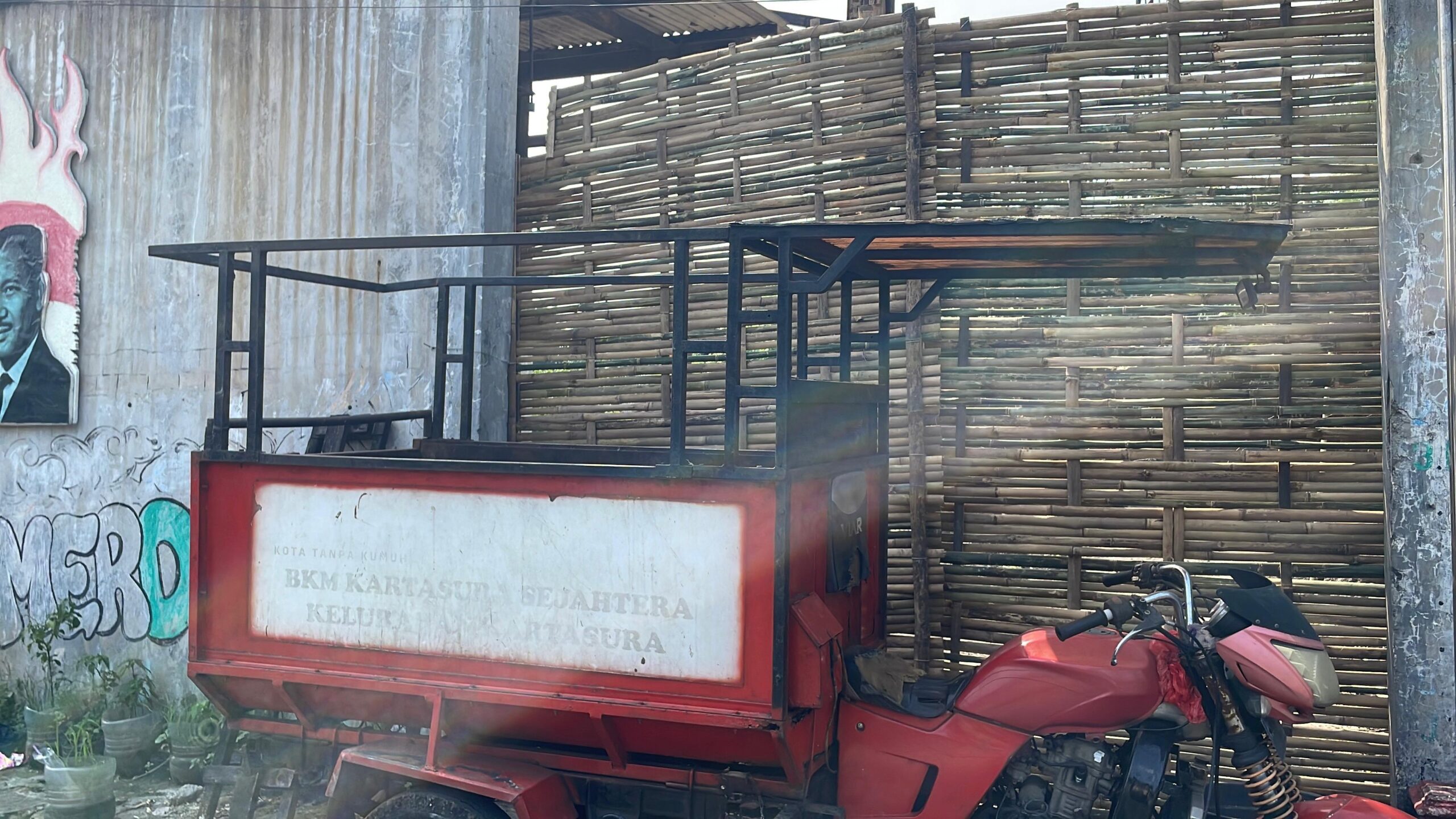 Red trailer with a metal frame and faded white panel parked beside a tall woven bamboo fence and graffiti wall on the left