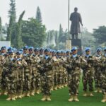 Rows of soldiers in camouflage uniforms and blue berets stand in formation on a grassy field rifles across chests with a statue in the background