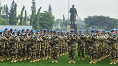 Rows of soldiers in camouflage uniforms and blue berets stand in formation on a grassy field rifles across chests with a statue in the background