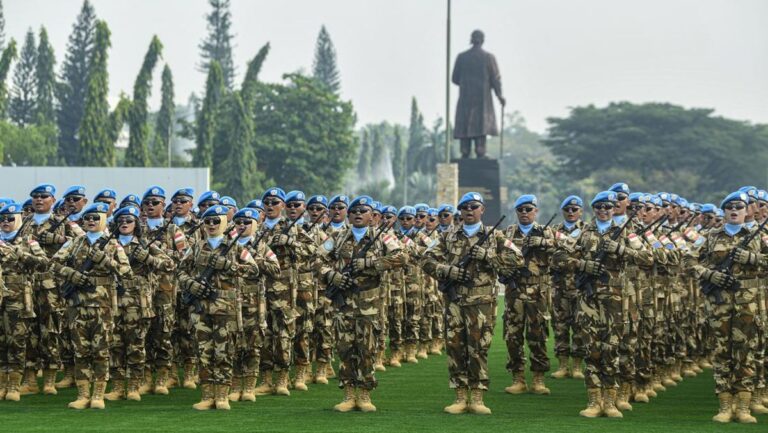 Rows of soldiers in camouflage uniforms and blue berets stand in formation on a grassy field, rifles across chests, with a statue in the background.