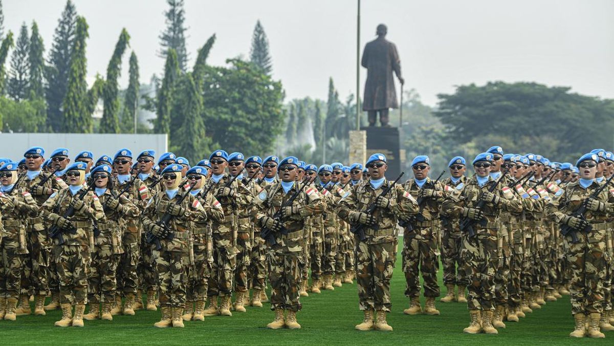 Rows of soldiers in camouflage uniforms and blue berets stand in formation on a grassy field rifles across chests with a statue in the background