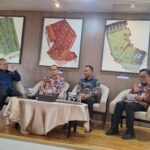 Panel discussion with four men seated on a stage one speaking into a microphone framed fabric art on the wall behind them