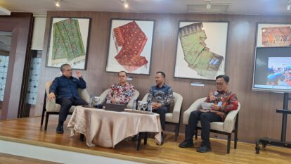 Panel discussion with four men seated on a stage, one speaking into a microphone, framed fabric art on the wall behind them.