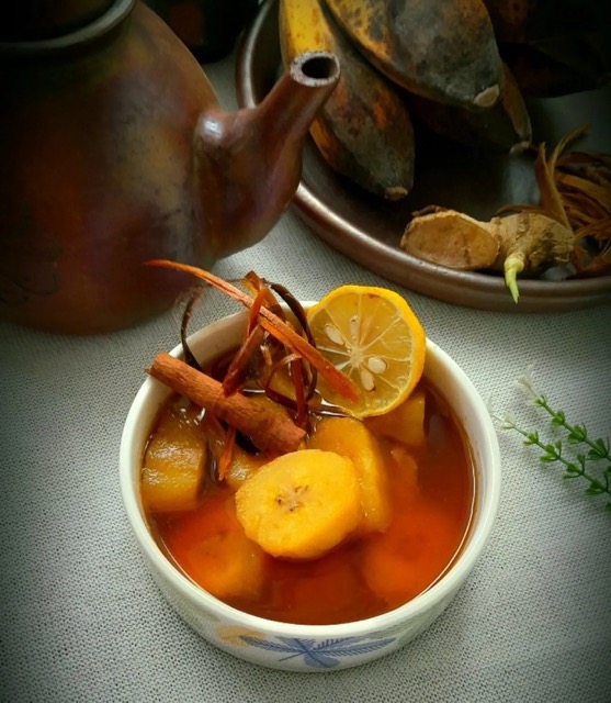 Cup of warm orange tea with orange slices cinnamon sticks and a lemon wheel set beside a teapot on a gray tablecloth