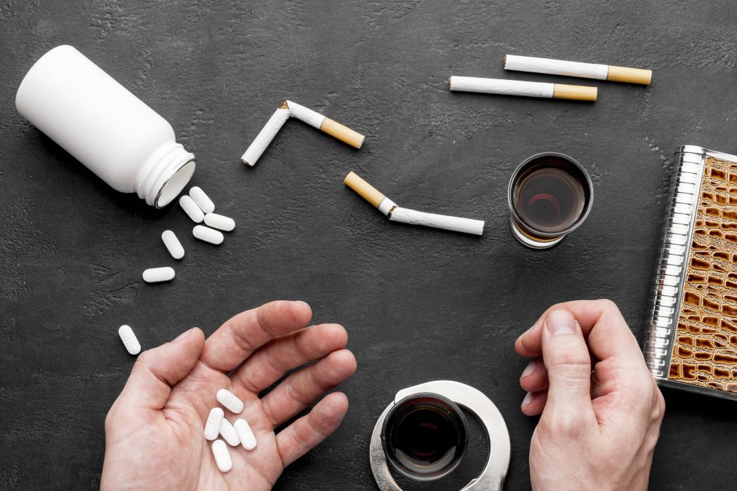 10fobat catrawarta Hands near a spilling white pill bottle with several pills on a dark surface surrounded by broken cigarettes a glass of dark liquid and a spiral notebookconveying an addiction related scene