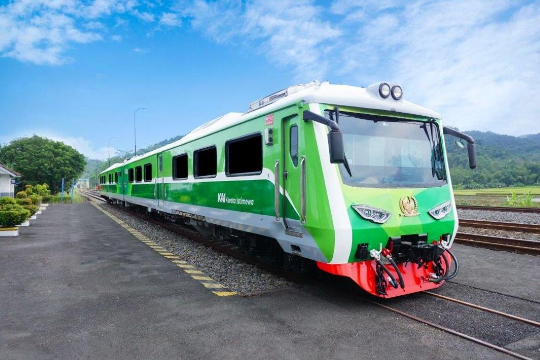 Green passenger train at a platform beside railway tracks on a sunny day with countryside in the background