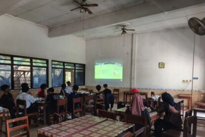 Group of students seated at tables in a classroom watching a soccer match projected on the wall