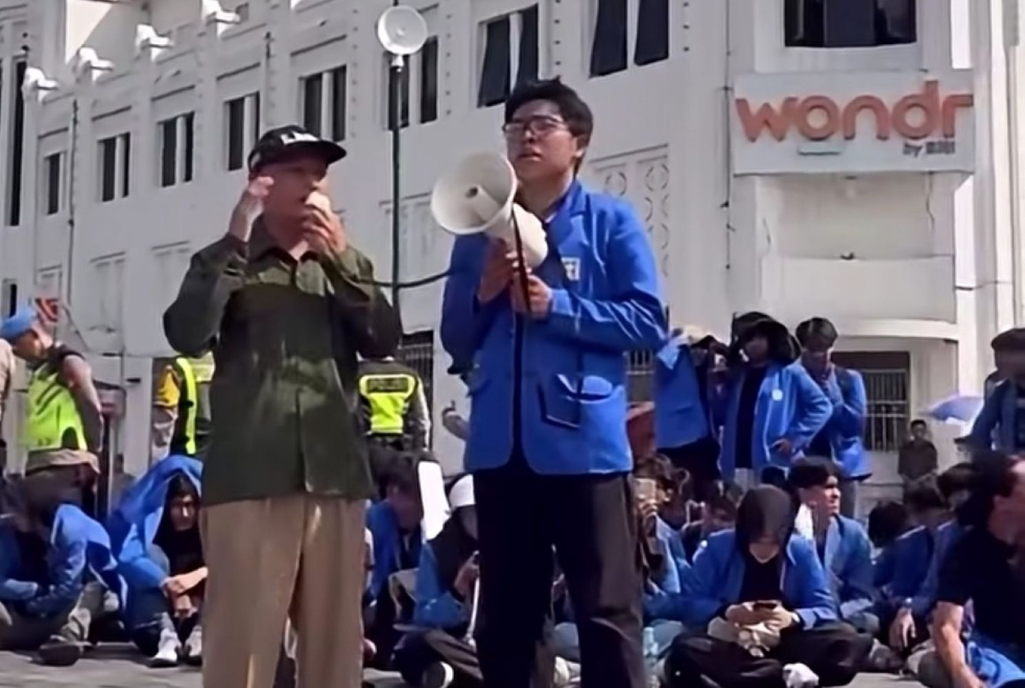 15fdemo catrawarta Man in blue jacket speaks into a megaphone while a man in olive green listens during a street protest with a seated crowd in blue jackets behind them