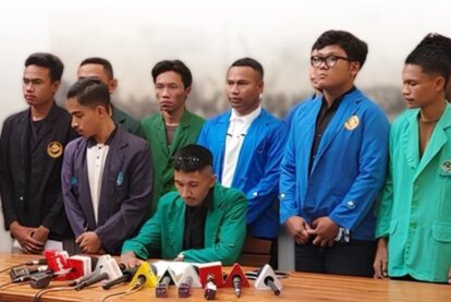 Man in a green blazer signs a document at a press conference while eight colleagues in colorful jackets stand behind him, microphones on the table.