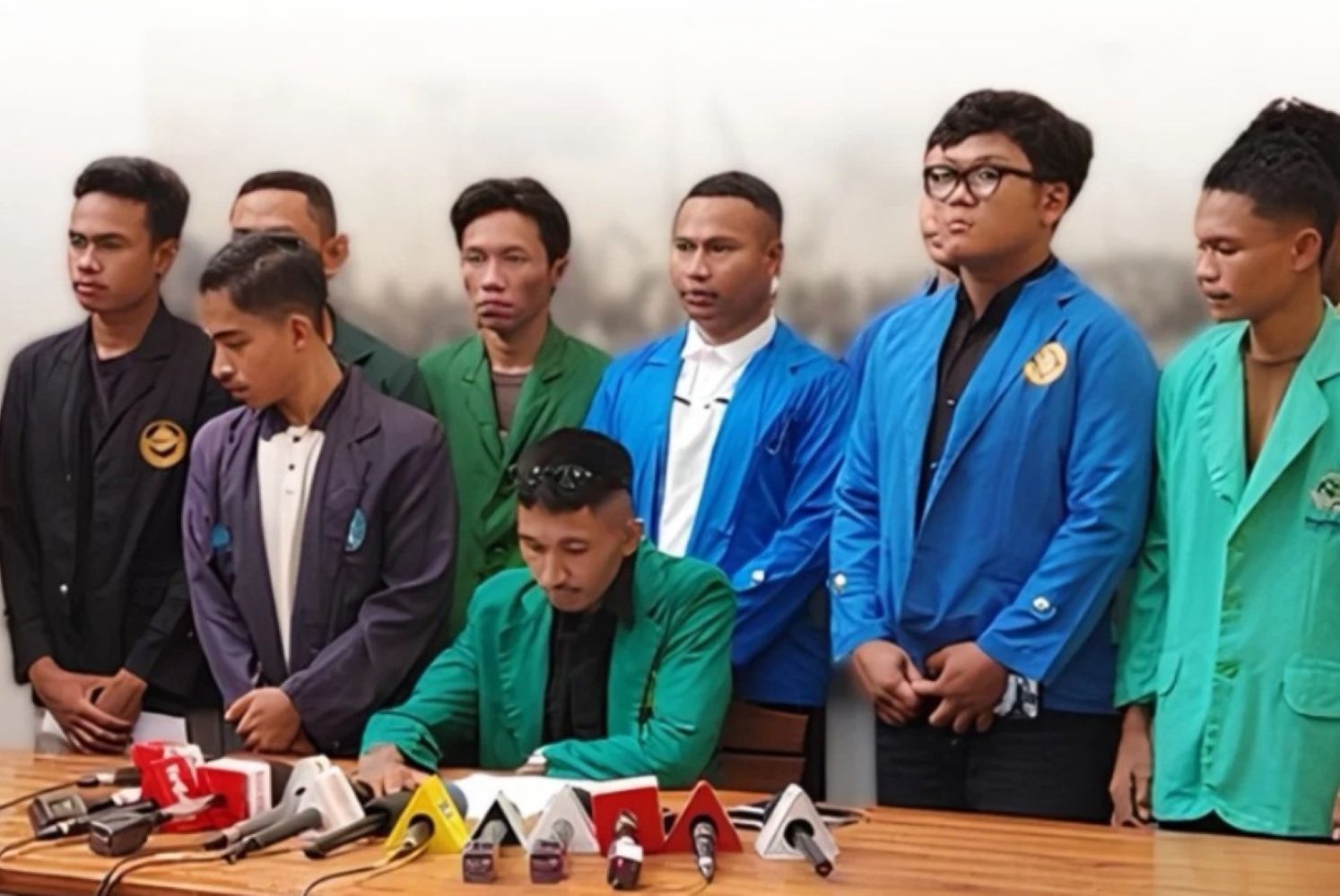 17fbem catrawarta Man in a green blazer signs a document at a press conference while eight colleagues in colorful jackets stand behind him microphones on the table