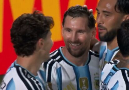 Two soccer players in Argentina national team jerseys celebrate together on the field, smiles visible amid a group celebration.