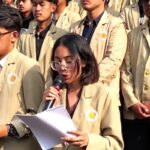 Young woman with glasses speaks into a microphone while reading papers surrounded by classmates in beige blazers at a school event