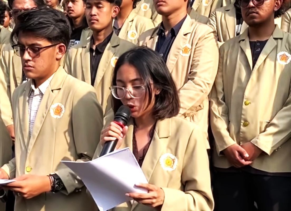 17fugm catrawarta Young woman with glasses speaks into a microphone while reading papers surrounded by classmates in beige blazers at a school event