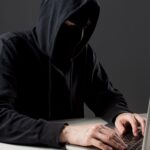 Person in a dark hoodie typing on a laptop face obscured by the hood in a dim setting