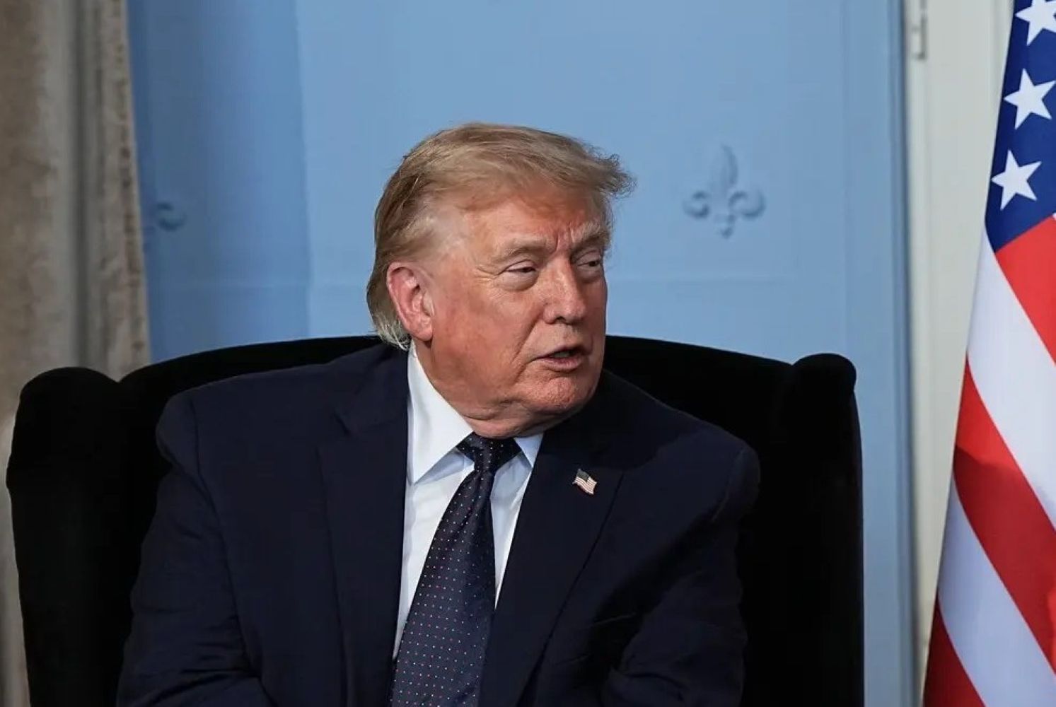 18ftrump catrawarta Older man in a dark suit and tie sits in a chair with part of a us flag visible to the right