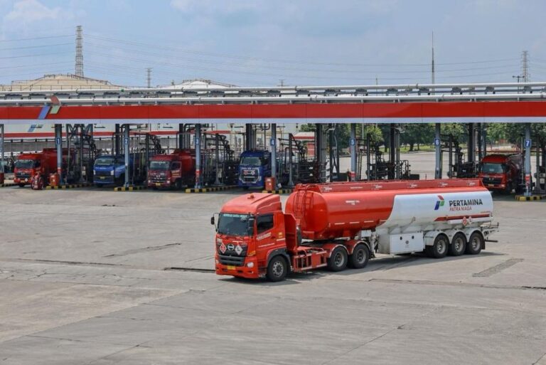 Red fuel tanker truck with a white trailer in the foreground at a Pertamina fuel depot; multiple loading bays and other trucks visible in the background.
