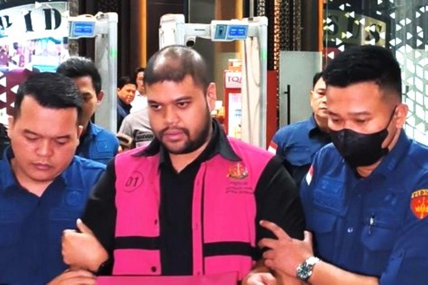19ftersangka catrawarta Man in a bright pink vest being escorted by two uniformed officers through a public area