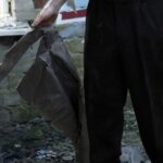 Person in black pants stands among rubble gripping a torn dark metal sheet blood on the right hand