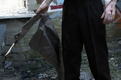 Person in black pants stands among rubble gripping a torn dark metal sheet blood on the right hand