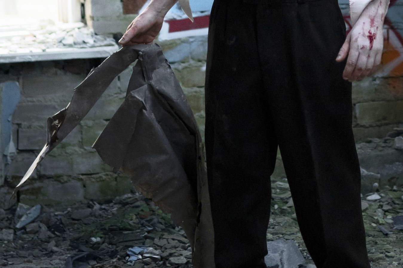 Person in black pants stands among rubble gripping a torn dark metal sheet blood on the right hand