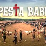 Festival banner reading pesta babi above a village square as people carry a pig carcass on a pole in a long line of spectators and participants