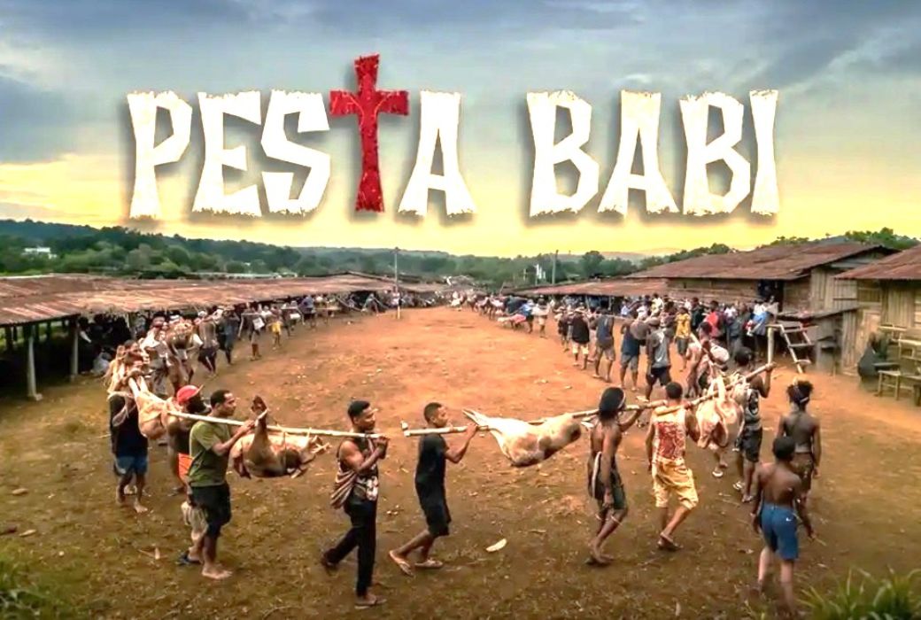 1fpesta catrawarta Festival banner reading pesta babi above a village square as people carry a pig carcass on a pole in a long line of spectators and participants