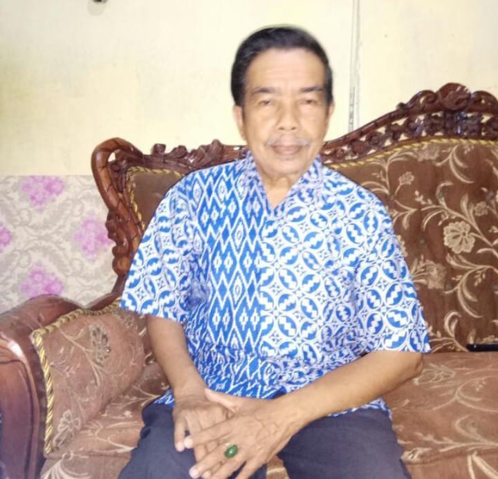 Older man seated on an ornate wooden sofa wearing a blue patterned shirt in a living room setting