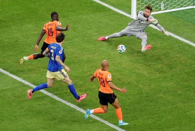 Soccer scene: orange defenders and a blue team player advance toward the goal while the goalie dives to save the ball near the net on a green pitch.