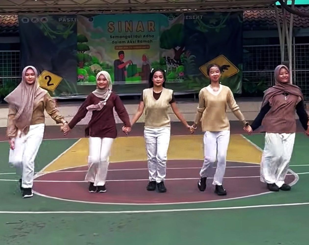21frukun1 catrawarta Five young women hold hands in a circle on a gymnasium basketball court during a performance wearing beige and brown outfits and white pants with headscarves or hijabs