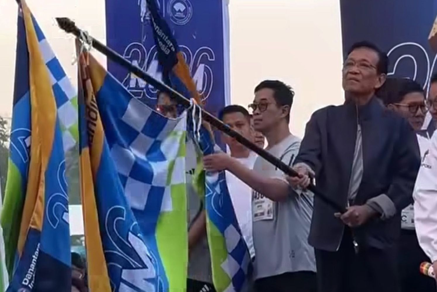 Group of people at a flag raising ceremony a man in a suit holds a flagpole while others wave blue and checkered flags in the background