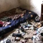 A cluttered dirty bedroom with a mattress on the floor and piles of trash and belongings scattered across the room