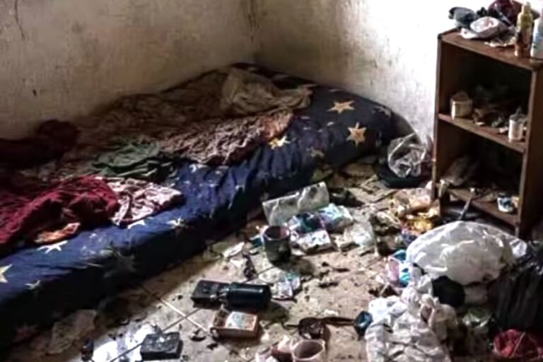 A cluttered, dirty bedroom with a mattress on the floor and piles of trash and belongings scattered across the room.