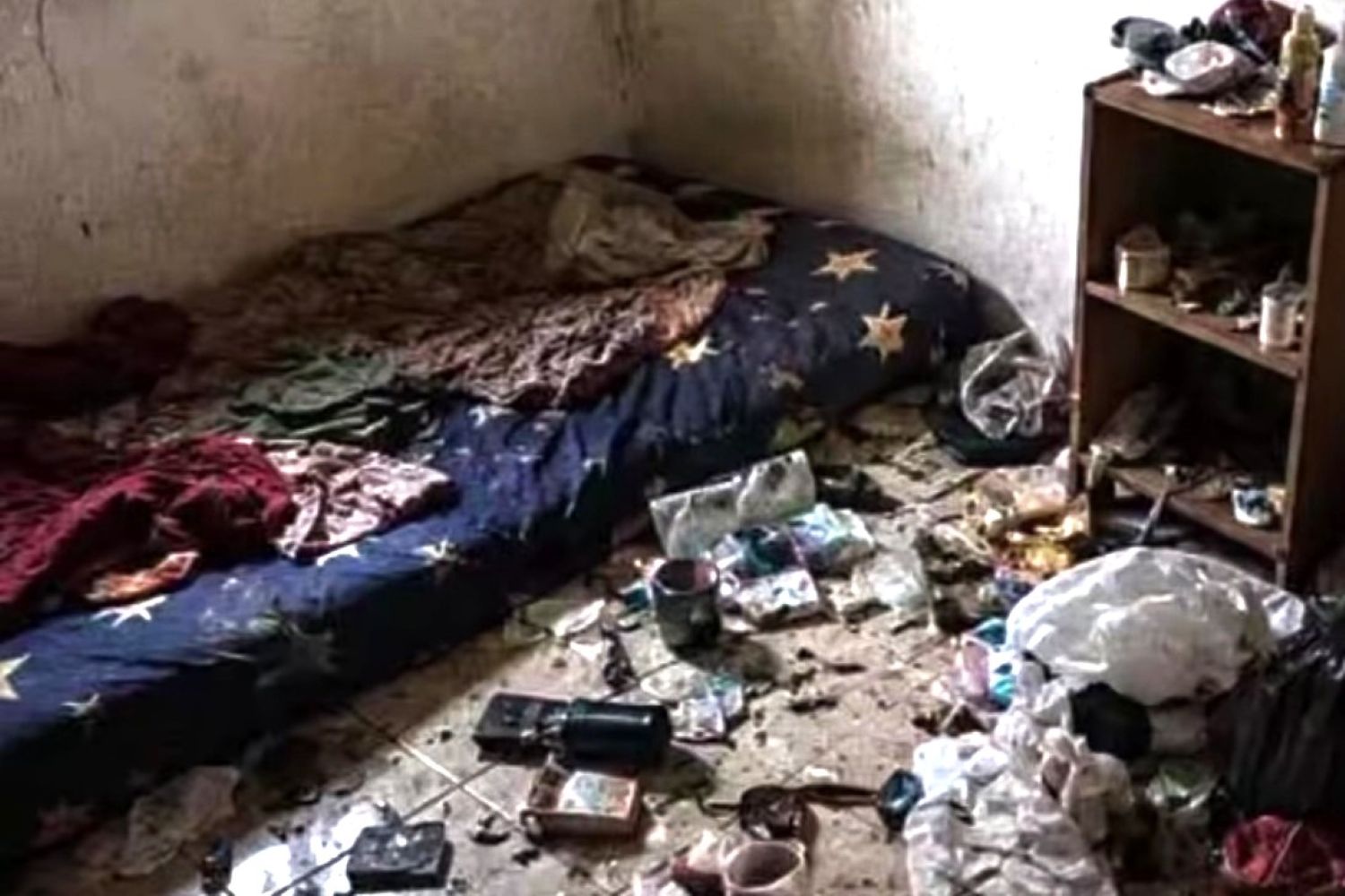 A cluttered dirty bedroom with a mattress on the floor and piles of trash and belongings scattered across the room