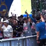 Crowd of runners behind a metal barrier celebrating at a race finish with confetti in the air