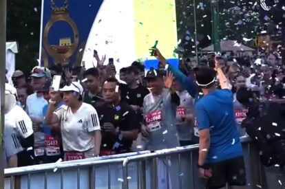 Crowd of runners behind a metal barrier celebrating at a race finish with confetti in the air.