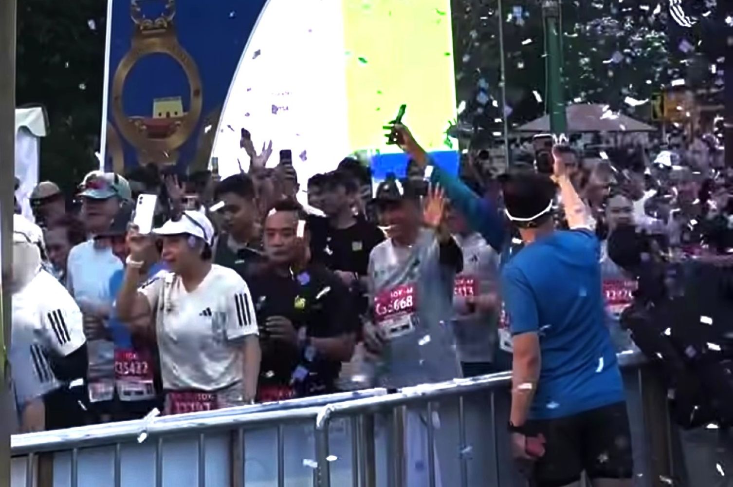 Crowd of runners behind a metal barrier celebrating at a race finish with confetti in the air