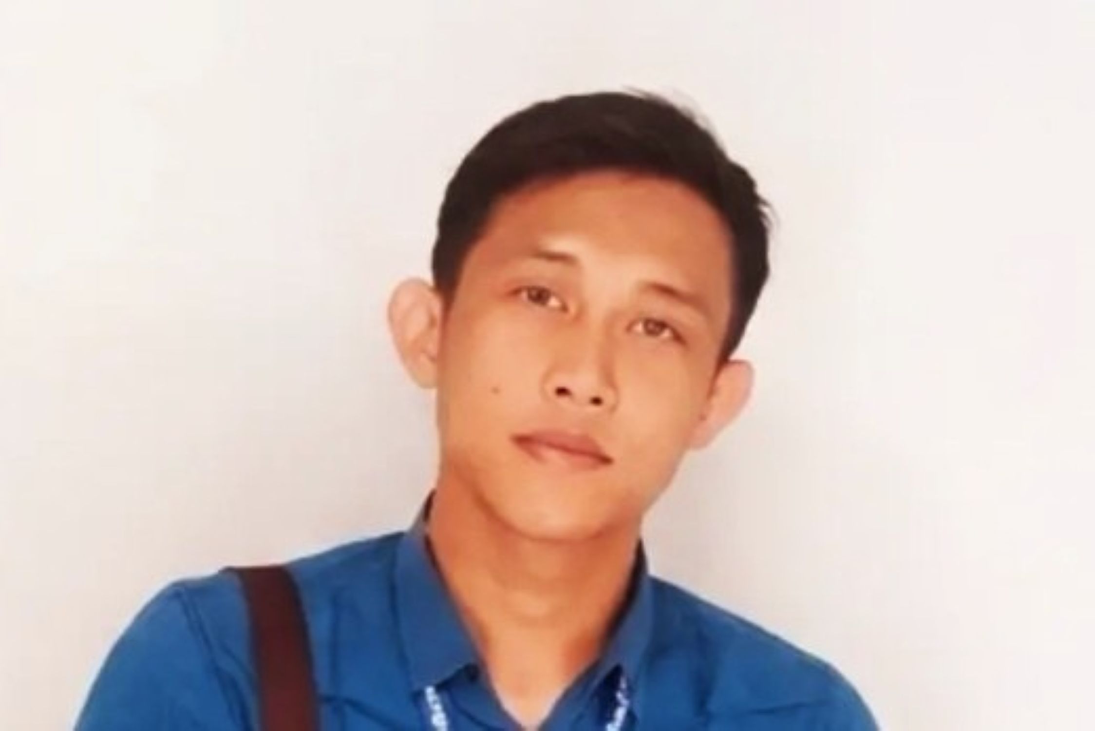 Portrait of a young man with short black hair wearing a blue shirt and a dark strap over his shoulder against a light background