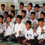 Group of indonesian students in white shirts and black caps sitting on a carpet and cheering one holds a microphone
