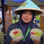 Woman in a blue hijab and conical hat at an outdoor event presenting two bowls with colorful fruit toppings and smiling
