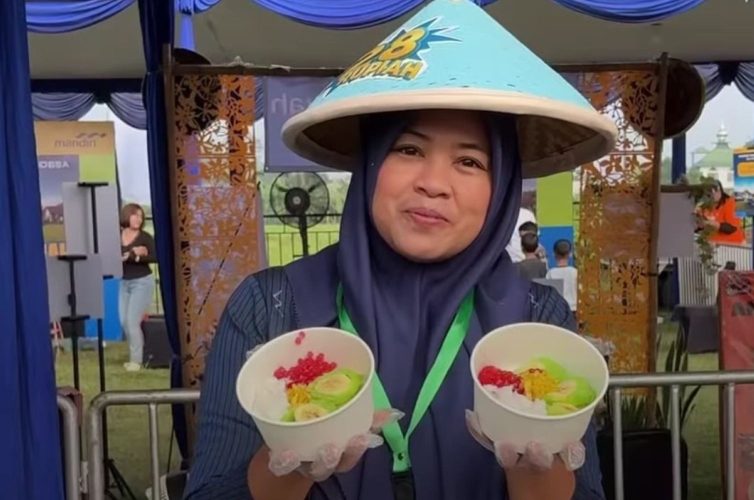 Woman in a blue hijab and conical hat at an outdoor event presenting two bowls with colorful fruit toppings and smiling