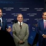 Man in a dark suit speaks into a microphone at a summit stage with a blue backdrop reading lake lucerne summit and flags behind