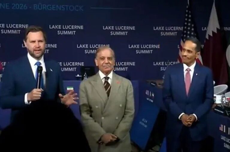 Man in a dark suit speaks into a microphone at a summit stage with a blue backdrop reading 'Lake Lucerne Summit' and flags behind.