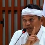 Man in white shirt and headband speaks into a microphone raising one finger in a gesture of emphasis in a formal setting
