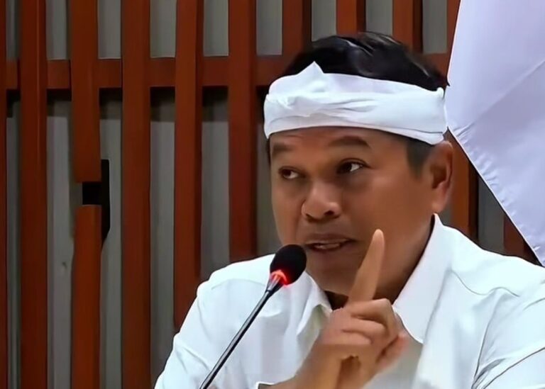 Man in white shirt and headband speaks into a microphone, raising one finger in a gesture of emphasis in a formal setting
