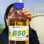 Person wearing a hijab holds up a large bottle with a blue cap labeled b50 biodiesel in a workshop setting